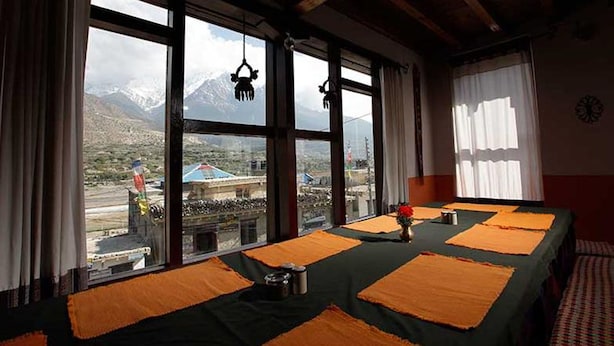 A room with large windows offering a mountain view, a table with orange placemats, and a clock on the wall. - Hotel Om's Home (Jomsom)