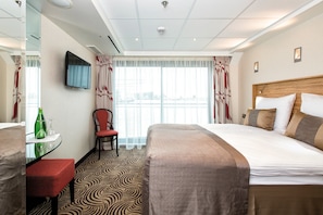 A hotel room with a bed, a desk with a chair, a television, and a view of the outside through large windows.