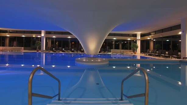 Piscina interior com um elemento central de água, rodeada por áreas de estar e grandes janelas.