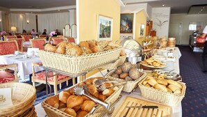 Disposição de buffet com pães e doces variados em cestos de vime, dispostos sobre uma mesa em área de refeições com cadeiras estofadas vermelhas e parede amarela ao fundo.