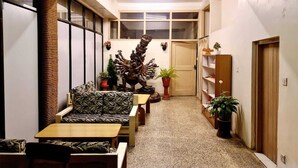 Un pasillo estrecho con un sofá, una mesa de madera y una escultura decorativa. Hay plantas en macetas y una puerta de madera al final.