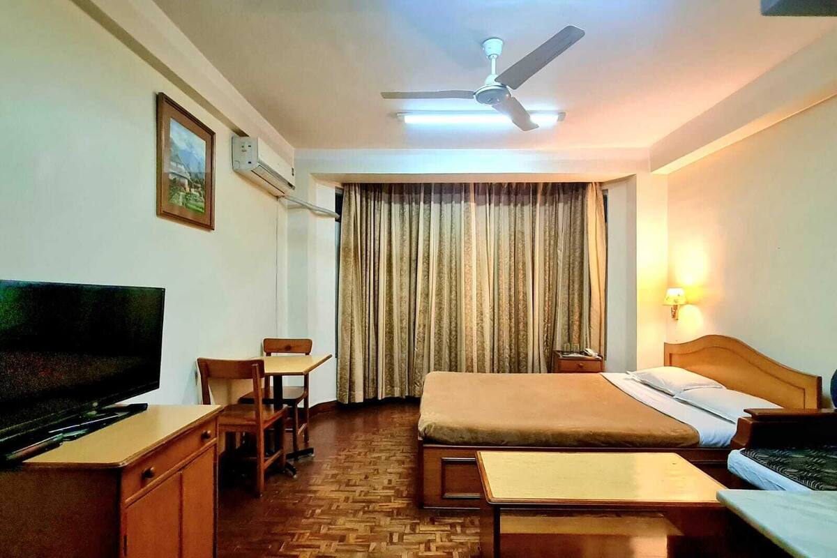 Habitació d'hotel amb un llit, una taula de treball amb una cadira, una televisió, un ventilador de sostre i una finestra amb cortines.