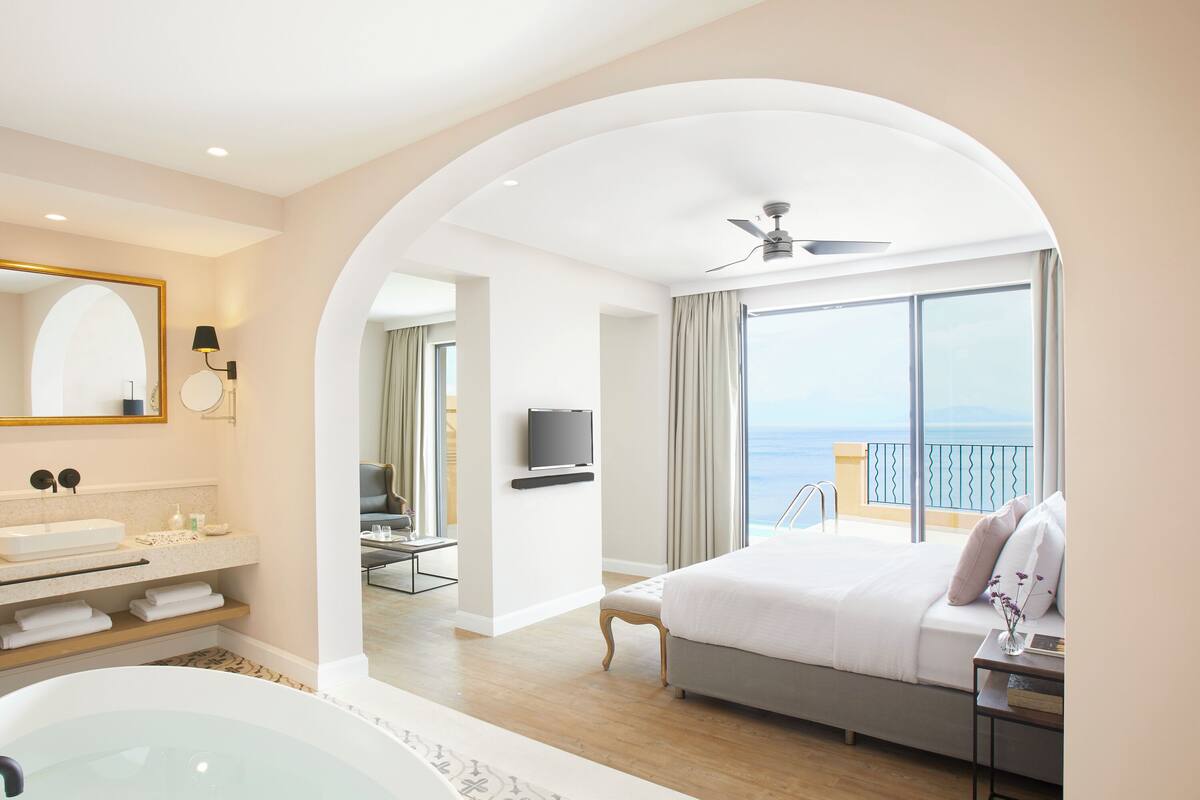 Una habitación de hotel moderna con una cama grande, un baño con bañera, televisión y un balcón con vistas al mar.