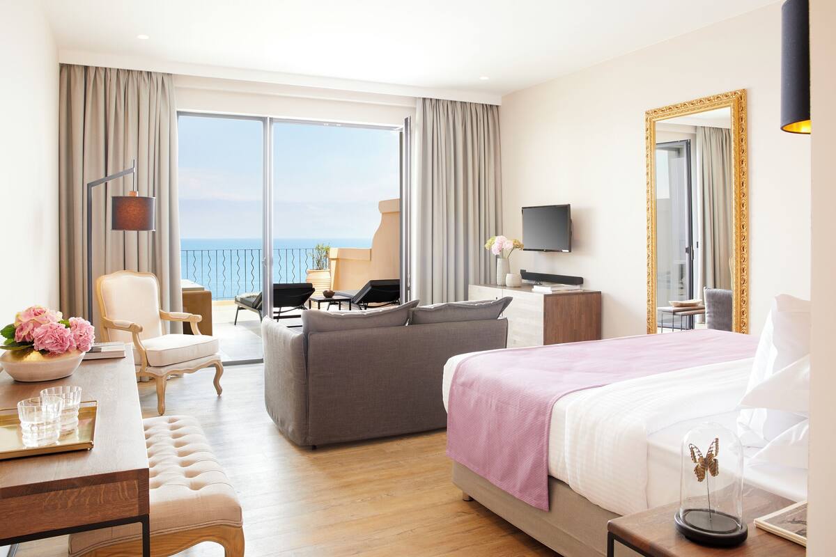 Habitación de hotel con una cama grande, un sofá, un escritorio con un florero y televisión, y vista al mar.