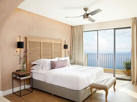 A bedroom with a large bed, a ceiling fan, a nightstand, a bedside table, a bench, and a view of the ocean through a balcony.