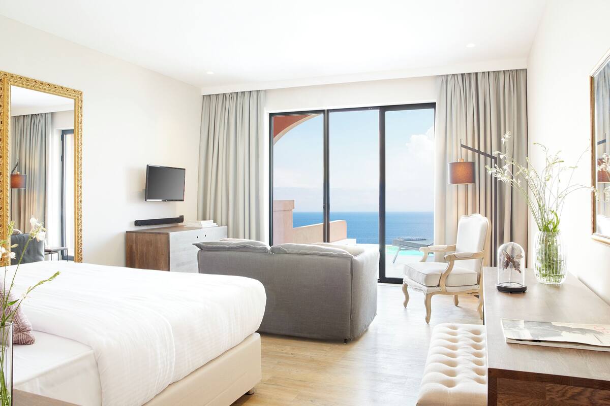 Habitación de hotel con una cama grande, un sofá gris, una mesita pequeña, una silla y vista al océano.