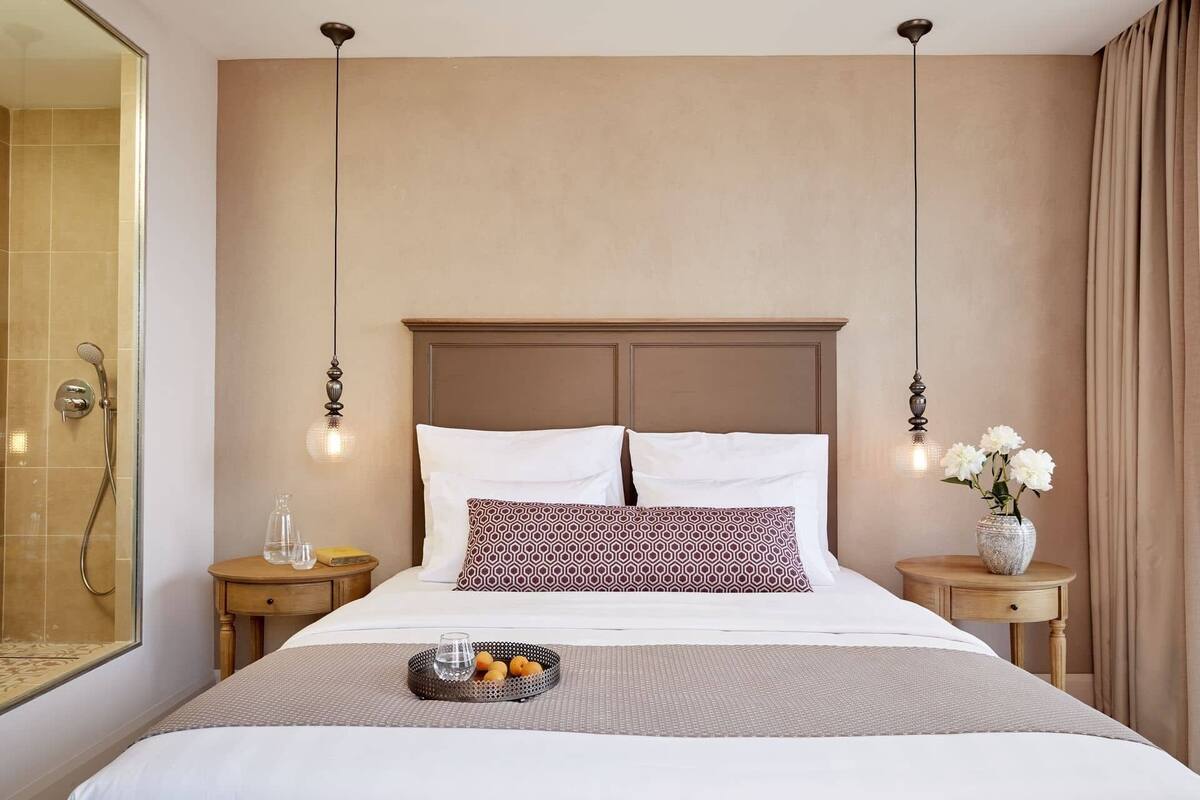 Habitación de hotel con una cama grande, dos mesitas de noche, zona de ducha y un florero con flores sobre una de las mesitas.