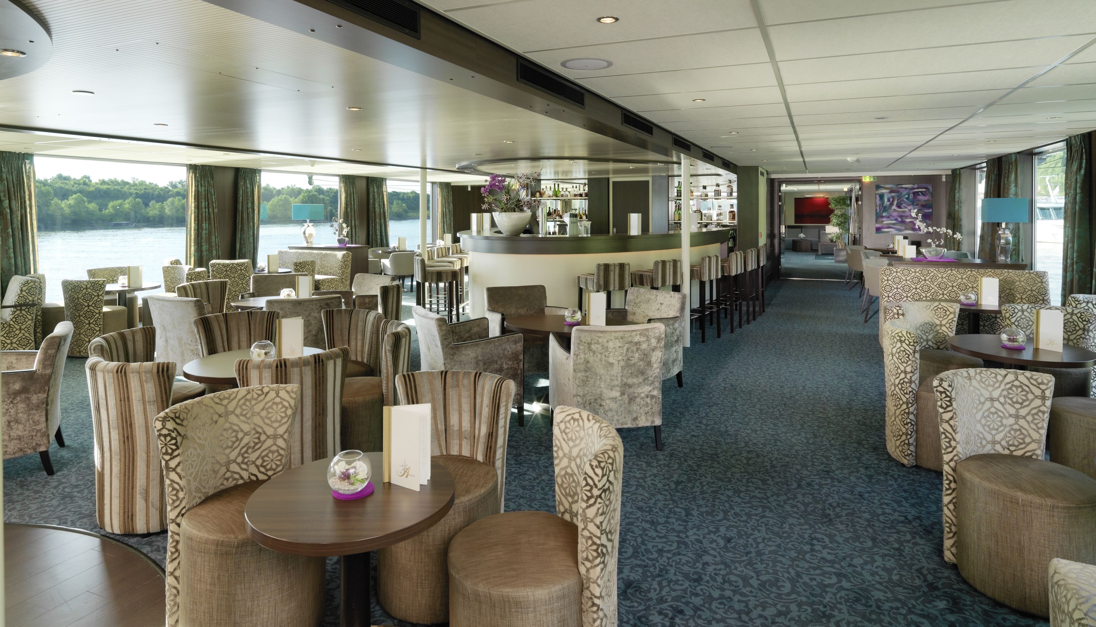 A spacious restaurant with a view of the water, featuring round tables and patterned chairs.