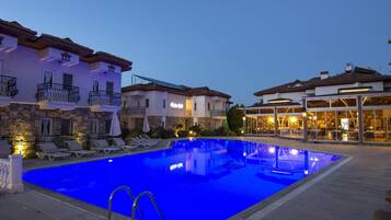 Una piscina con sillones de descanso, un edificio de fachada de piedra y una estructura con frentes de vidrio e iluminación cálida.