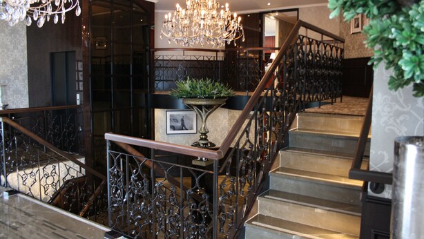 A grand staircase with ornate metal railings and a chandelier, leading to a landing with a potted plant and framed picture. - Crossgates Hotelship Messe - Düsseldorf (Düsseldorf)