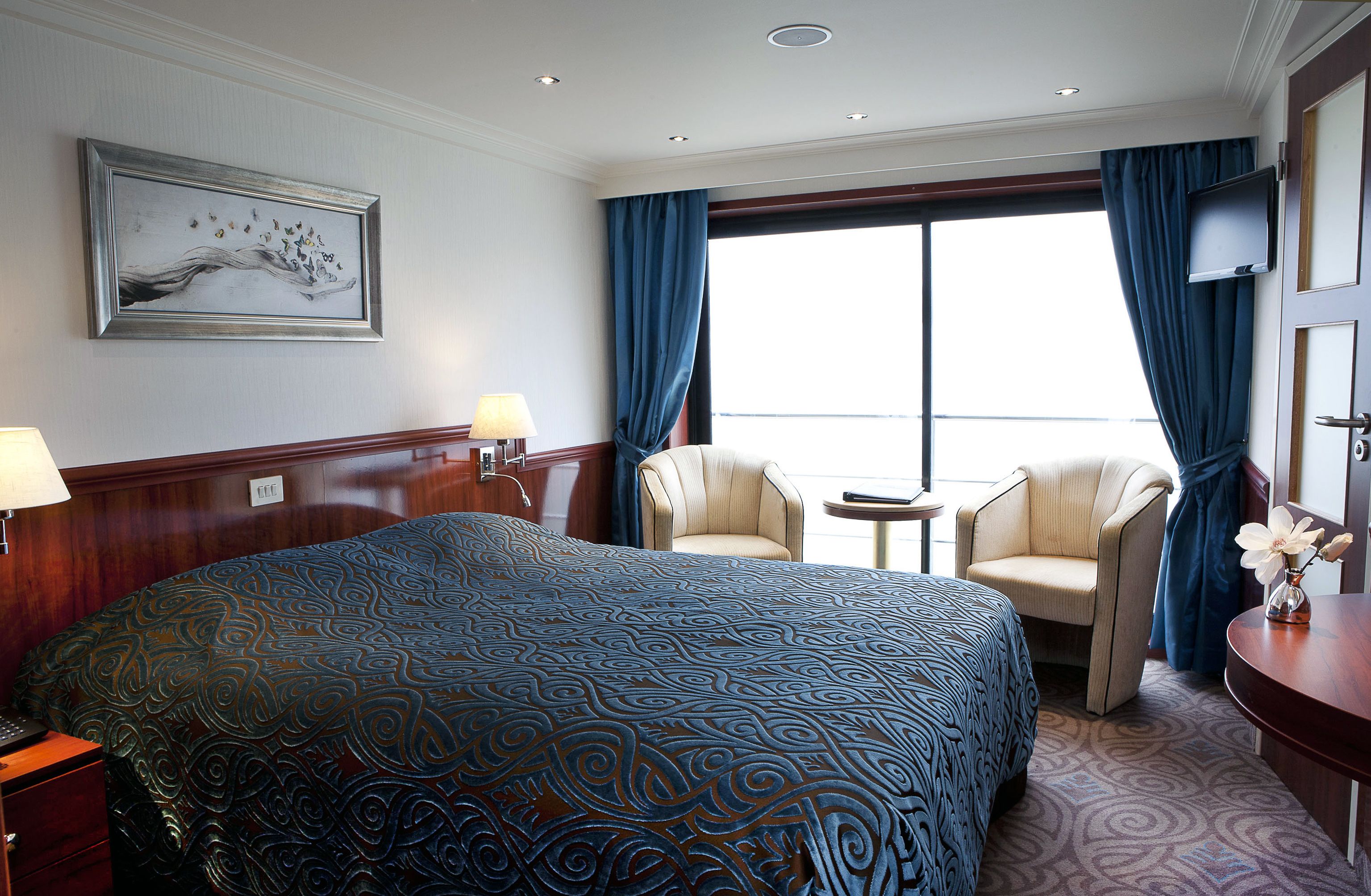 A hotel room with a large bed, two armchairs, a small table, a TV, and a window with curtains.
