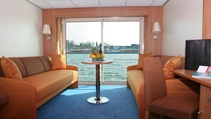 A room with two sofas, a small table with fruit and a bottle, a chair, a desk with a computer, and a large window with a view of the water. - Crossgates Hotelship Messe - Düsseldorf (Düsseldorf)