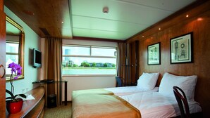 A hotel room with a large bed, a desk with a TV, a chair, and a window with a view of buildings and greenery. - Crossgates Hotelship Altstadt - Düsseldorf (Düsseldorf)