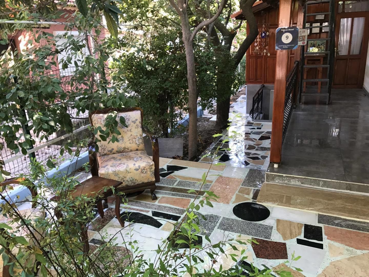 A tiled outdoor area with a chair and a bench, surrounded by trees and a building entrance.