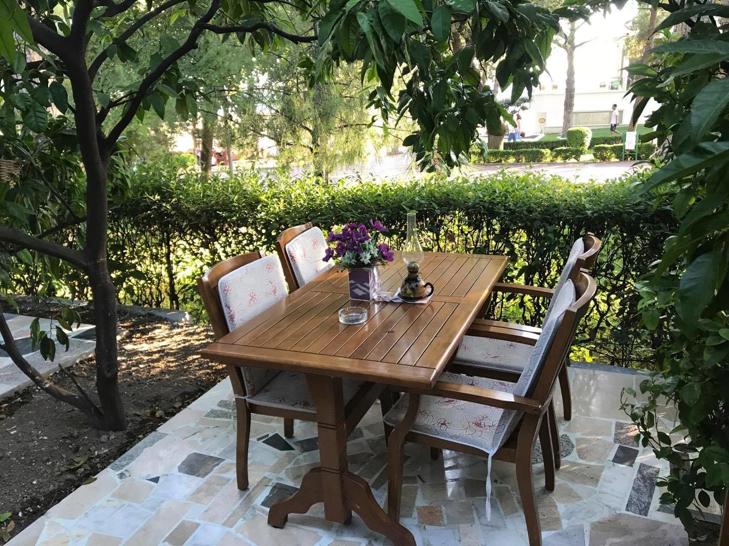 Uma mesa de jantar de madeira com cadeiras, rodeada por um jardim com sebes e árvores.