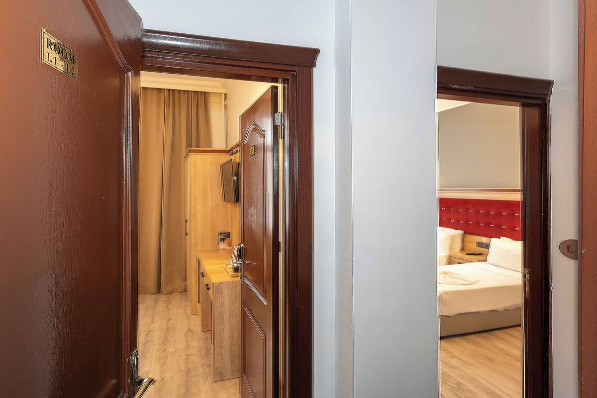 A hotel room with a wooden door, a bed with a red headboard, and a visible bathroom area with a towel rack.