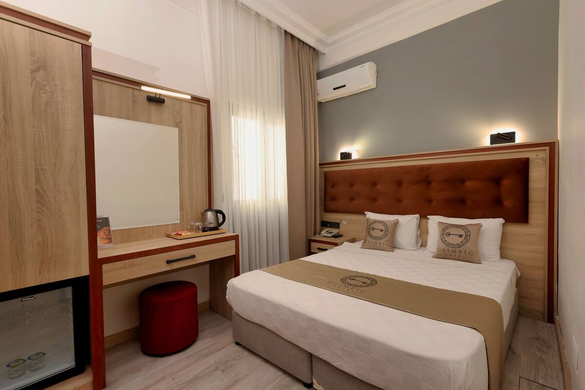 A hotel room with a large bed, a wooden headboard, a desk with a chair, a red waste bin, and a large mirror.