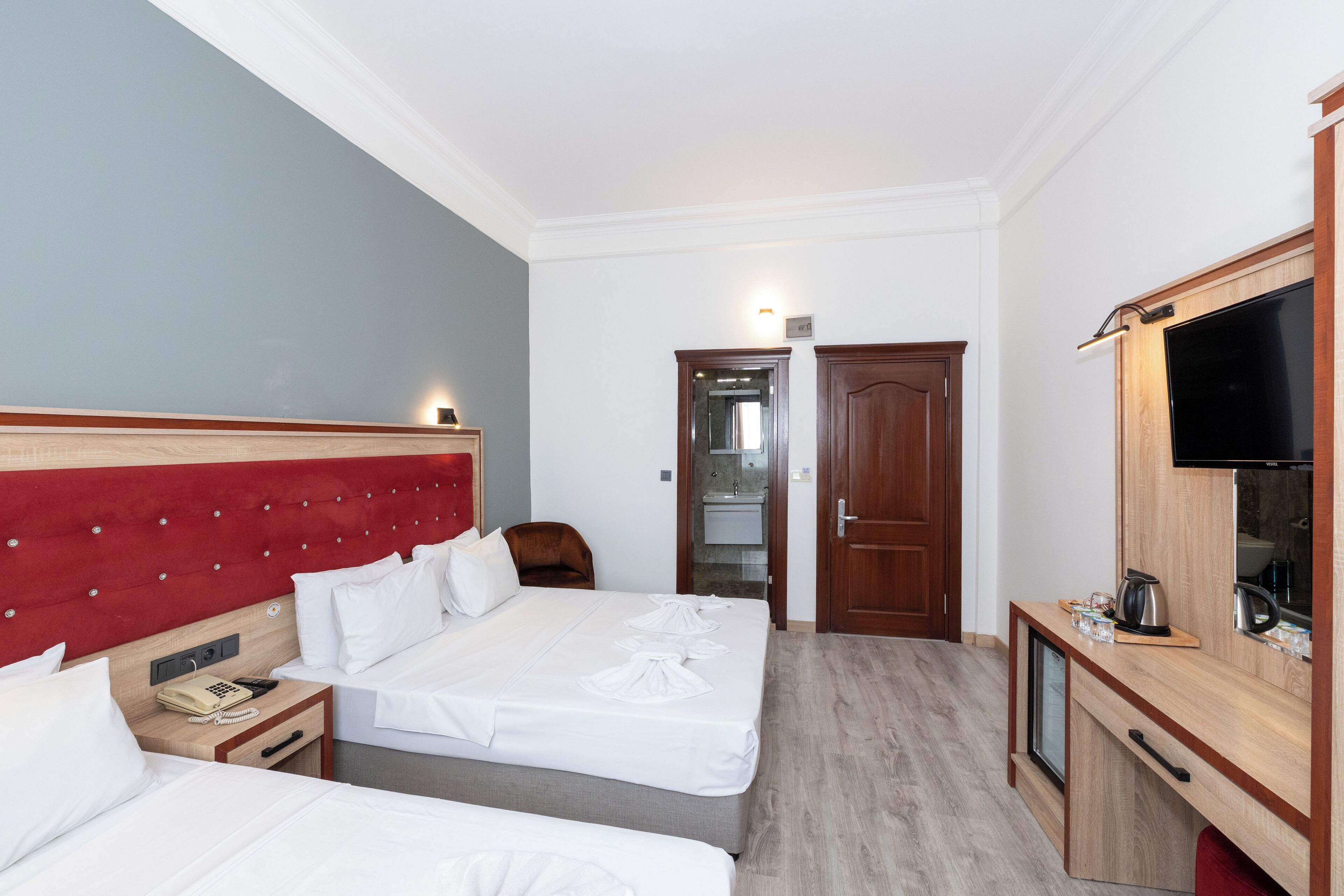 Quarto de hotel com duas camas, cabeceira de madeira, televisão, uma escrivaninha pequena e espelho.