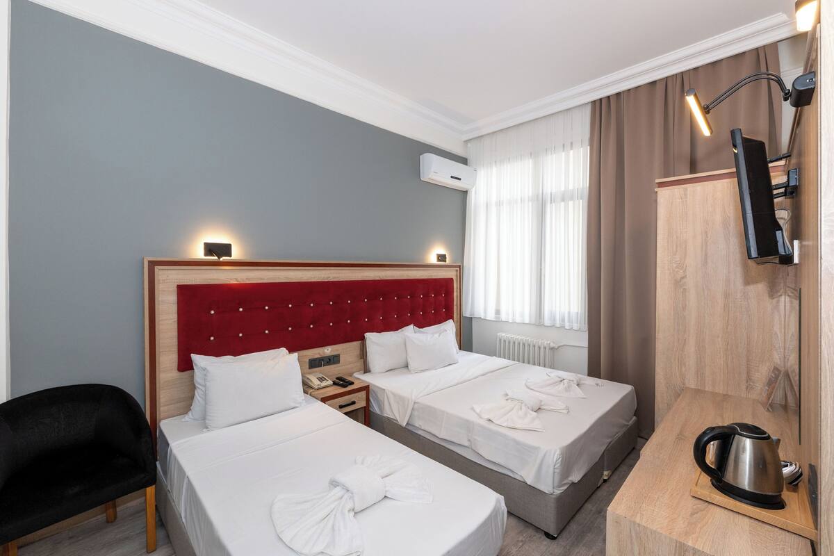 A hotel room with two beds, a red headboard, a wooden sideboard, a black chair, and a kettle.