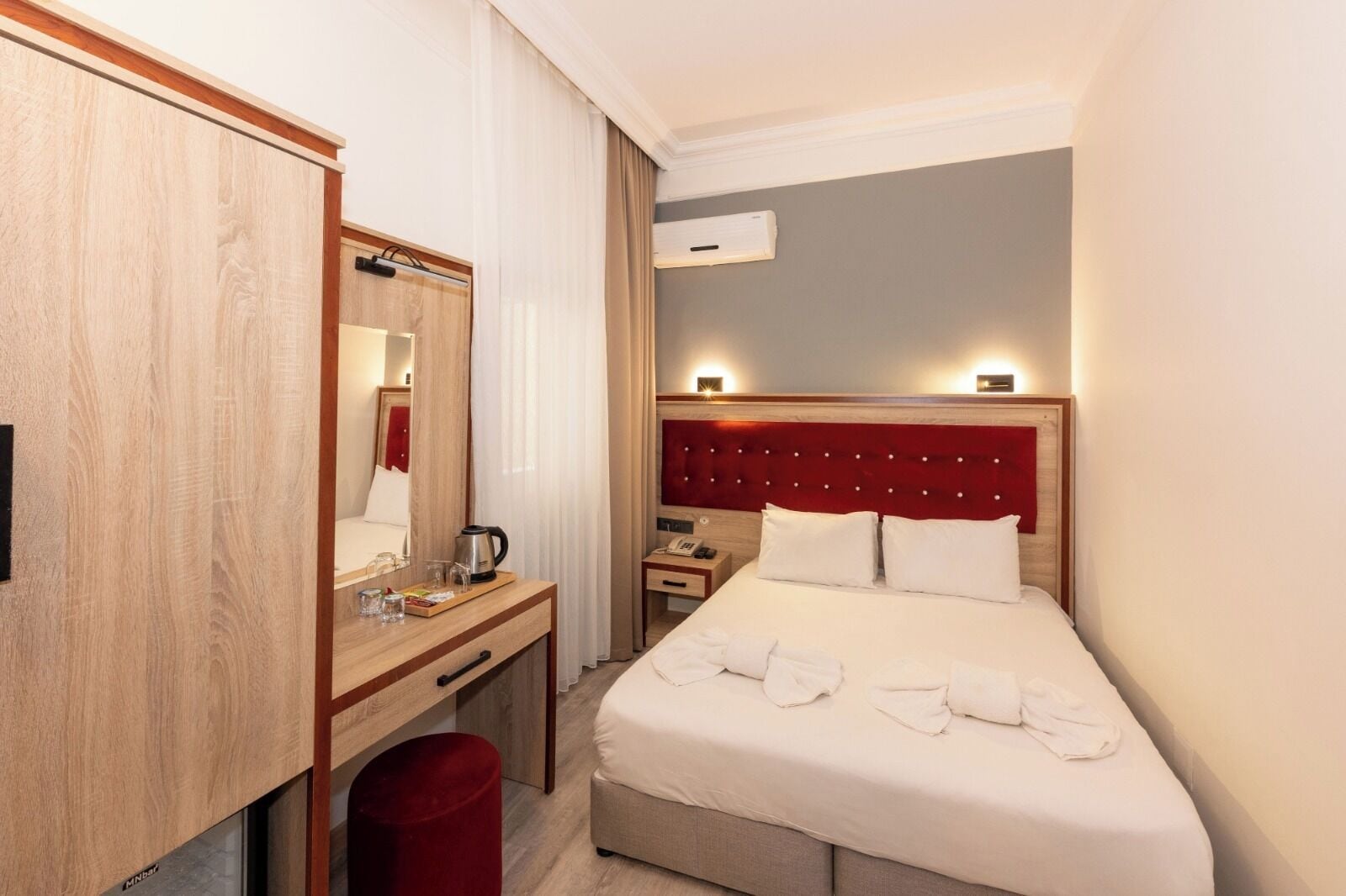 A hotel room with a bed, a desk, a mirror, a red trash bin, and an air conditioning unit.