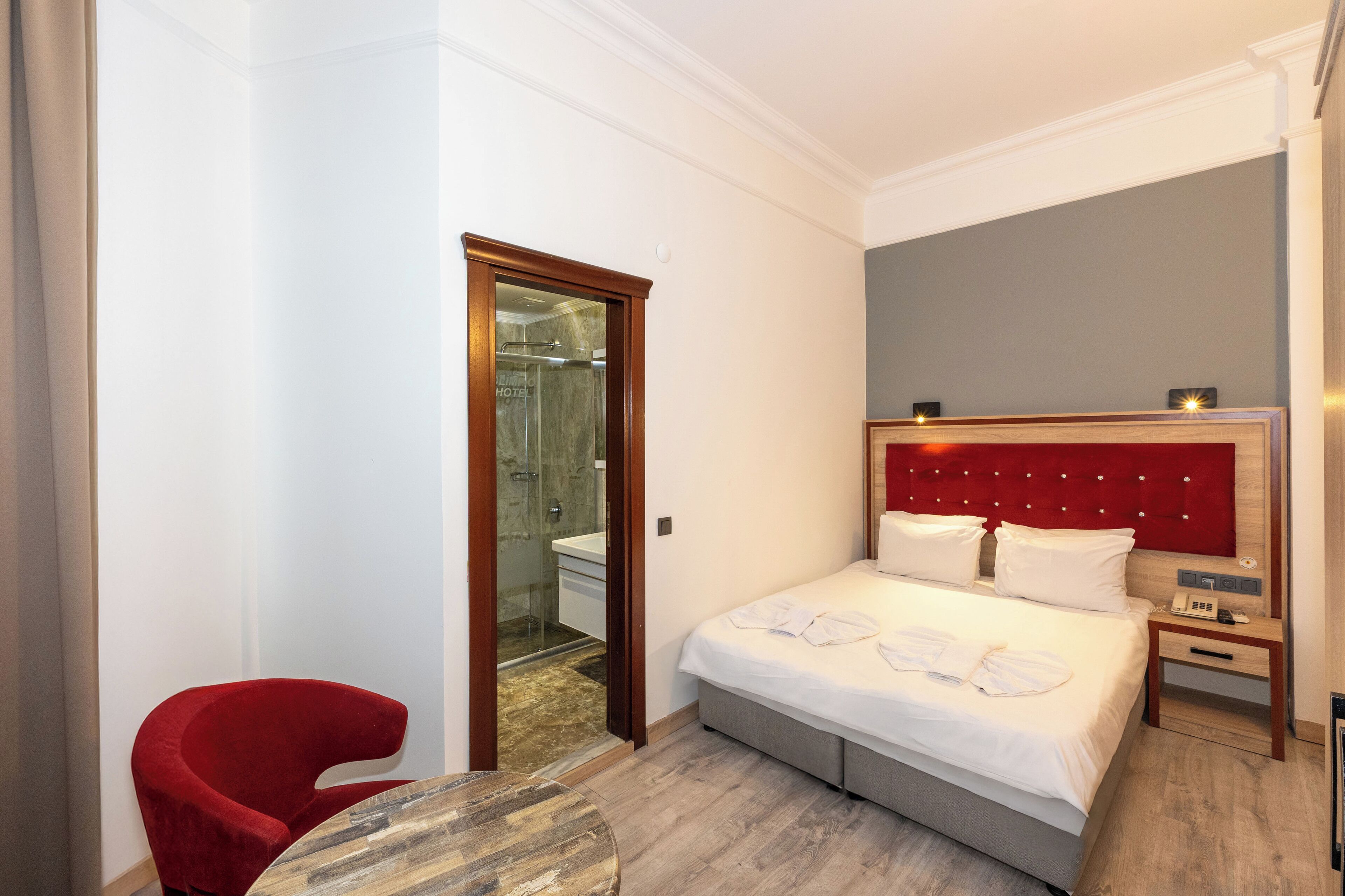 Een hotelkamer met een bed, een rode stoel, een houten tafel, een nachtkastje en een badkamer die door een geopende deur zichtbaar is.