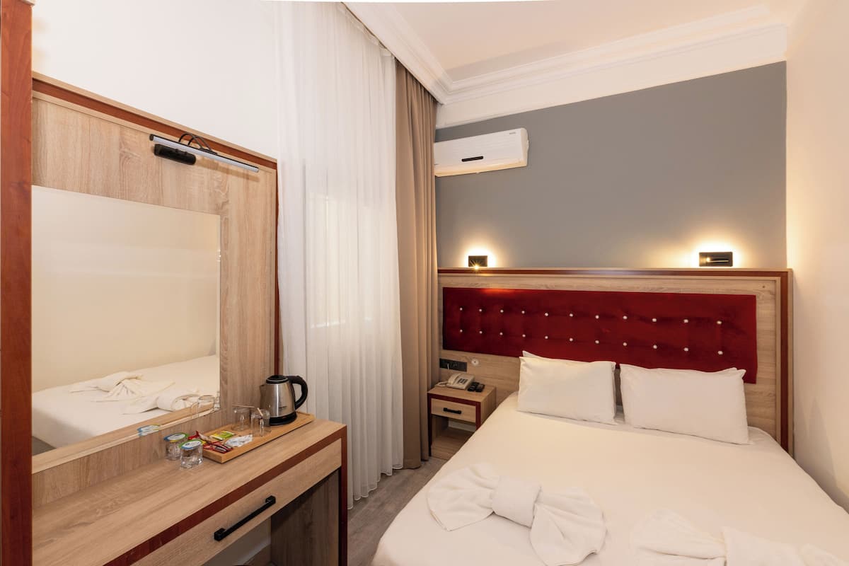 A hotel room with a large bed, a bedside table with a lamp, a mirror, a wooden nightstand, and an air conditioning unit.