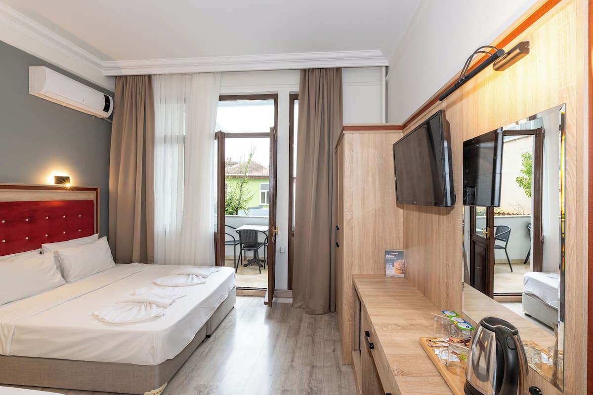 A hotel room with a bed, a desk with a kettle, a TV, and an air conditioner.