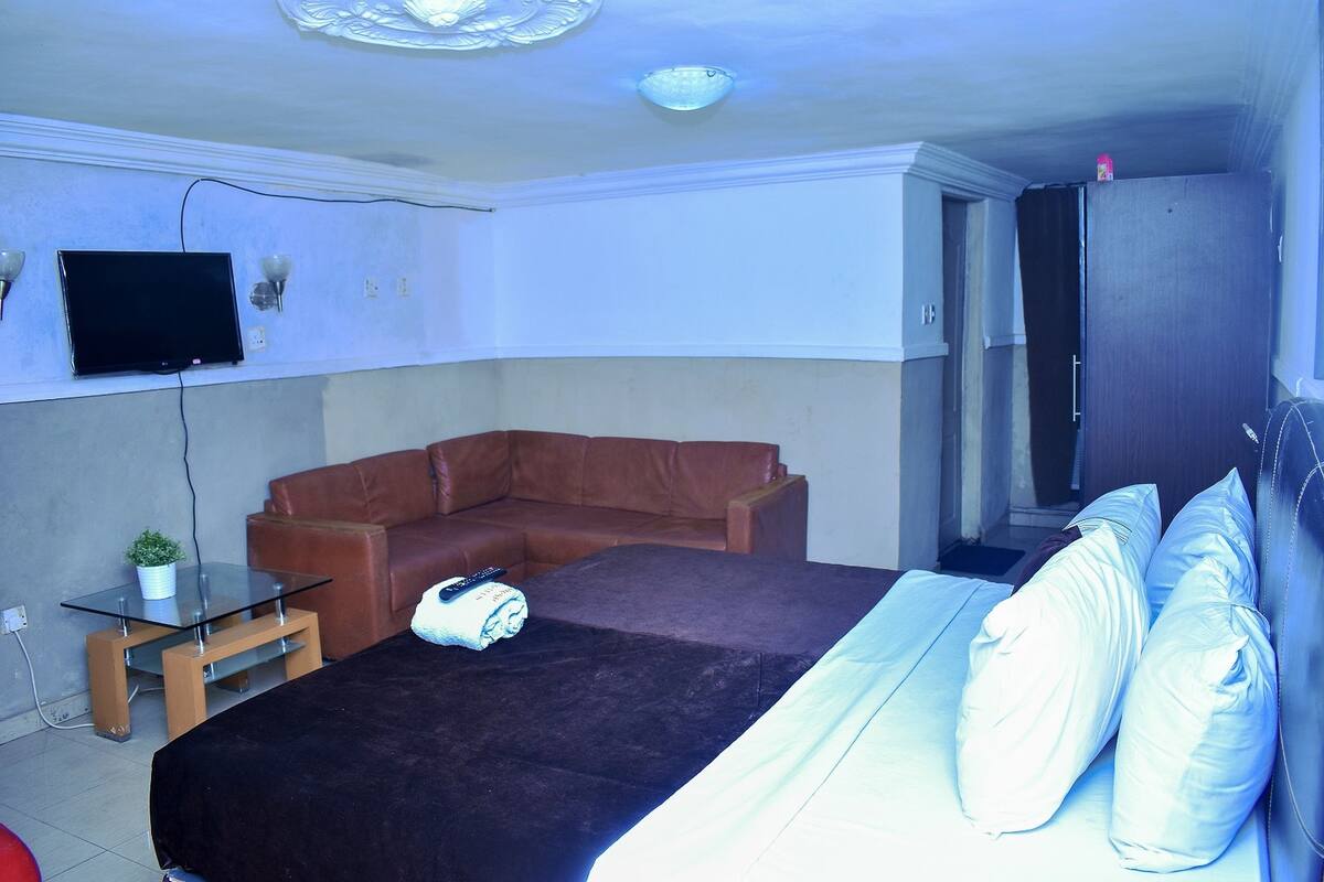 Une chambre avec un lit, un canapé en cuir marron, une table basse en verre, un téléviseur fixé au mur et une armoire.