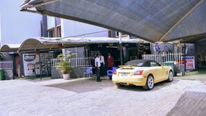 Ingresso di un hotel con una zona esterna coperta, un'auto decappottabile gialla parcheggiata nelle vicinanze e un cartello per una palestra.