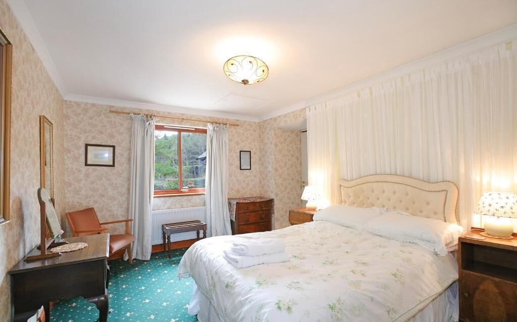 Un dormitorio con cama, escritorio, silla, ventana con cortinas y mesita de noche con lámpara.