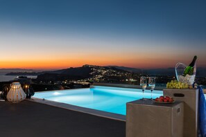 Une piscine sur le toit avec vue sur le coucher de soleil, deux verres de vin, une bouteille de champagne et un panier de fruits.