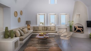 A modern living room with a sofa, a coffee table, and two wicker chairs. There are three windows with open shutters, a flat-screen TV mounted on the wall, and a fireplace with a decorative piece. - White & Co. Exclusive Island Villas (Santorini)