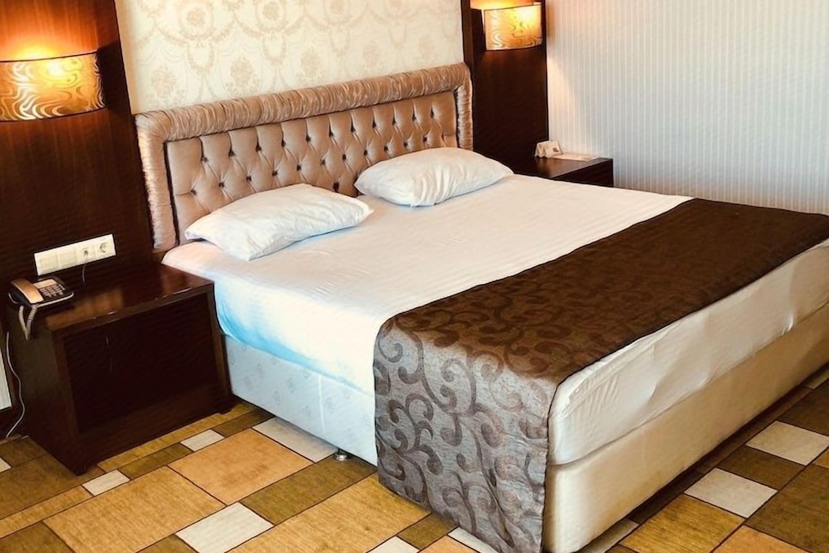 Una habitación de hotel con una cama grande, un cabecero con diseño estampado, dos lámparas de noche, una mesita de noche y un suelo a cuadros.