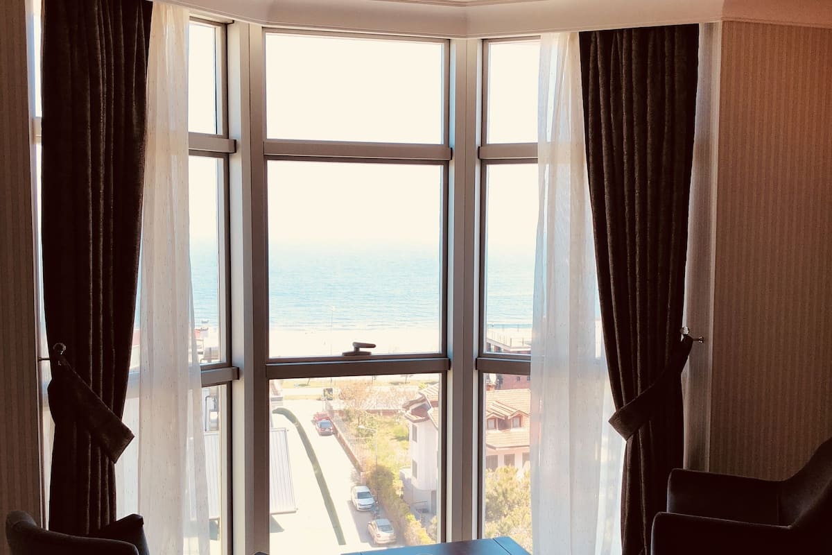 A room with a large bay window, two chairs, a small table, and a view of the sea and buildings.
