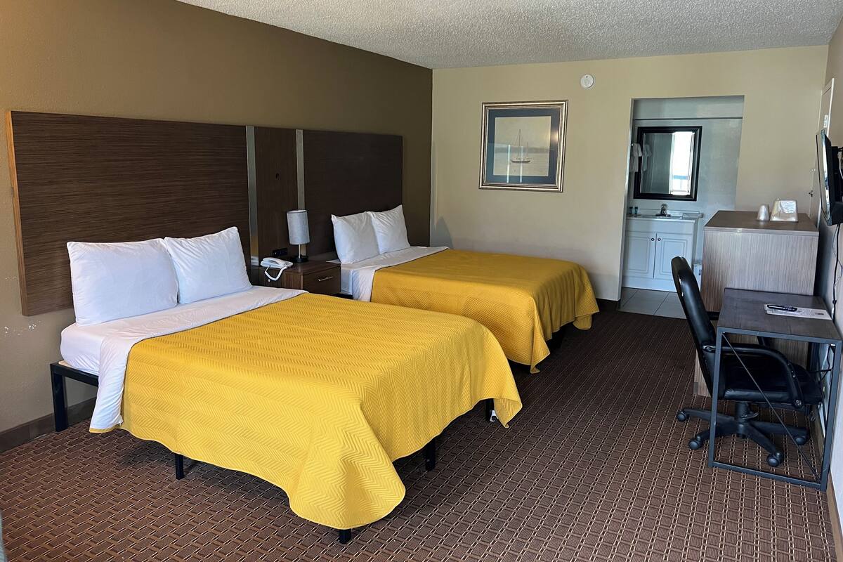 A hotel room with two beds, a desk, a chair, a mirror, and a small bathroom.