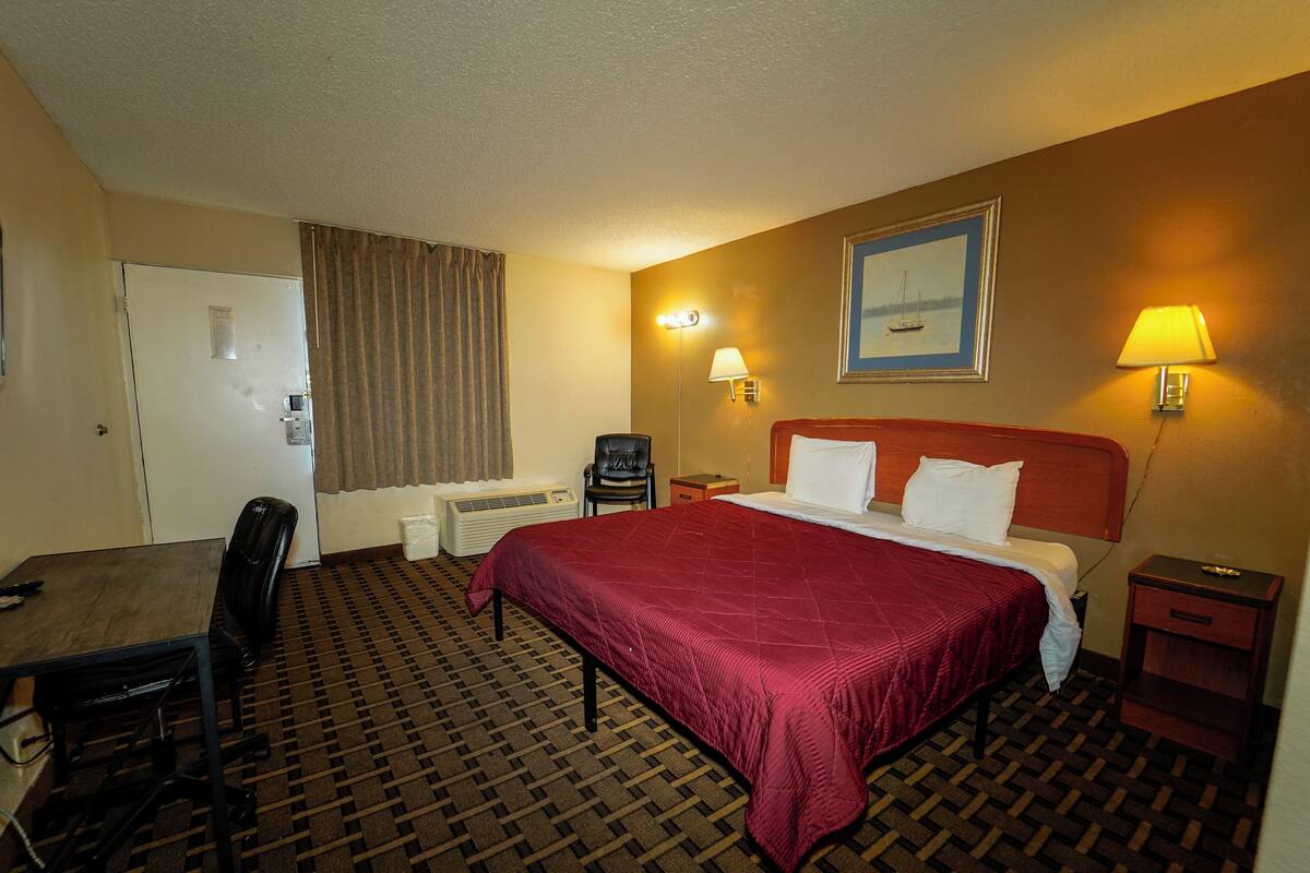 A hotel room with a bed, a desk, a chair, a nightstand, and a lamp.