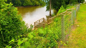 Eine Holzbrücke über einem ruhigen Fluss, umgeben von üppigem Grün und einem eingezäunten Bereich.