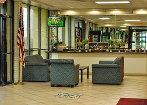 A reception area with two patterned armchairs, a small table, a welcome sign, and a television displaying a weather forecast. - Red Carpet Inn & Suites (Kinston)
