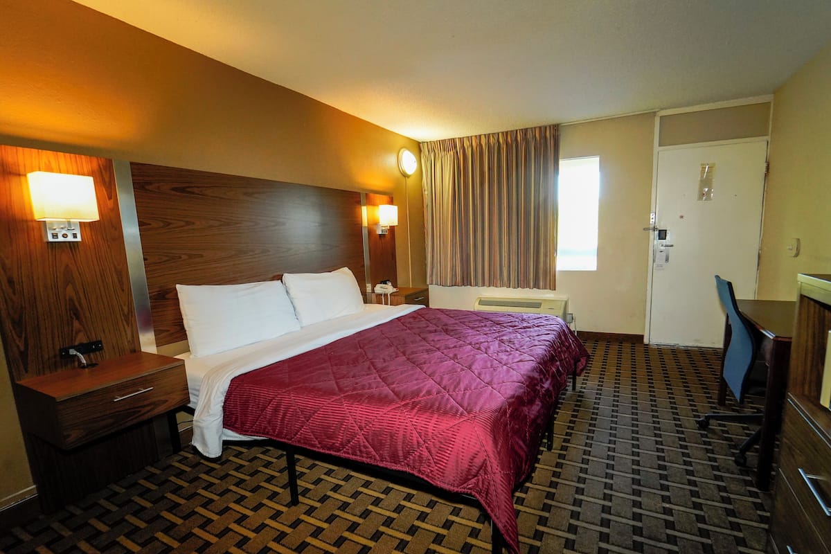 A hotel room with a bed, a nightstand, a desk, a chair, a window with curtains, and a door.
