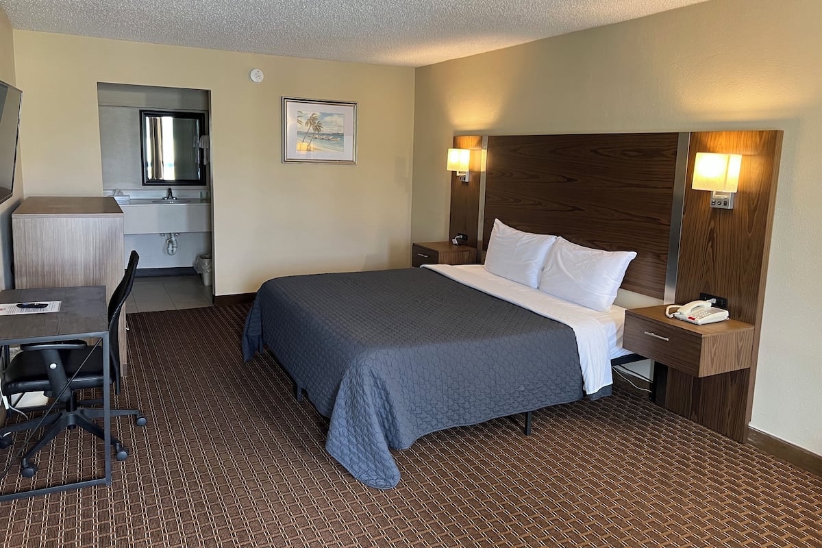 A hotel room with a bed, a desk, a chair, a nightstand, a lamp, and a painting on the wall.