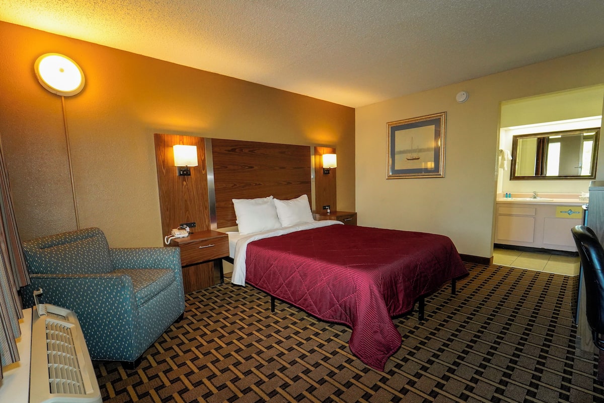 A hotel room with a bed, a desk with a chair, a small table, a nightstand, a lamp, a mirror, and a bathroom with a sink and mirror.