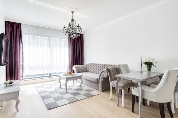 A modern living room with a grey sofa, a dining table with chairs, a chandelier, and a large window with curtains.