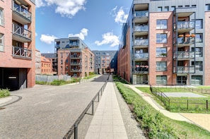 A modern urban street with multi-story residential buildings, a paved walkway, and a clear sky.