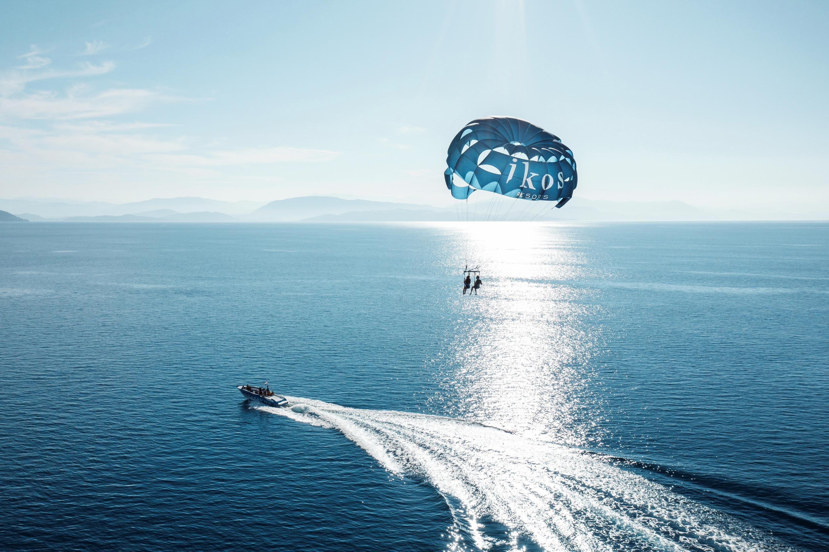 Un parapendio sopra un mare calmo, con un'imbarcazione che crea un'onda e una costa in lontananza.