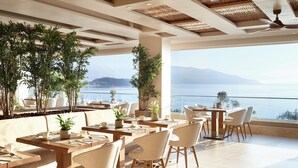 Un espacio moderno para comer al aire libre con varias mesas y sillas, rodeado de plantas en macetas, y vistas panorámicas a un lago y montañas lejanas.