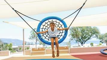 A child on a swing with a netted seat, surrounded by outdoor seating and a view of the sea.
