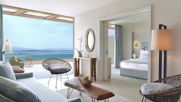 Una habitación de hotel moderna con balcón que da al mar, una cama, un área de estar con sofá y sillas, y una mesa de comedor.