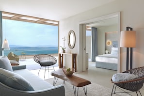 Una habitación de hotel moderna con balcón que da al mar, una cama, un área de estar con sofá y sillas, y una mesa de comedor.