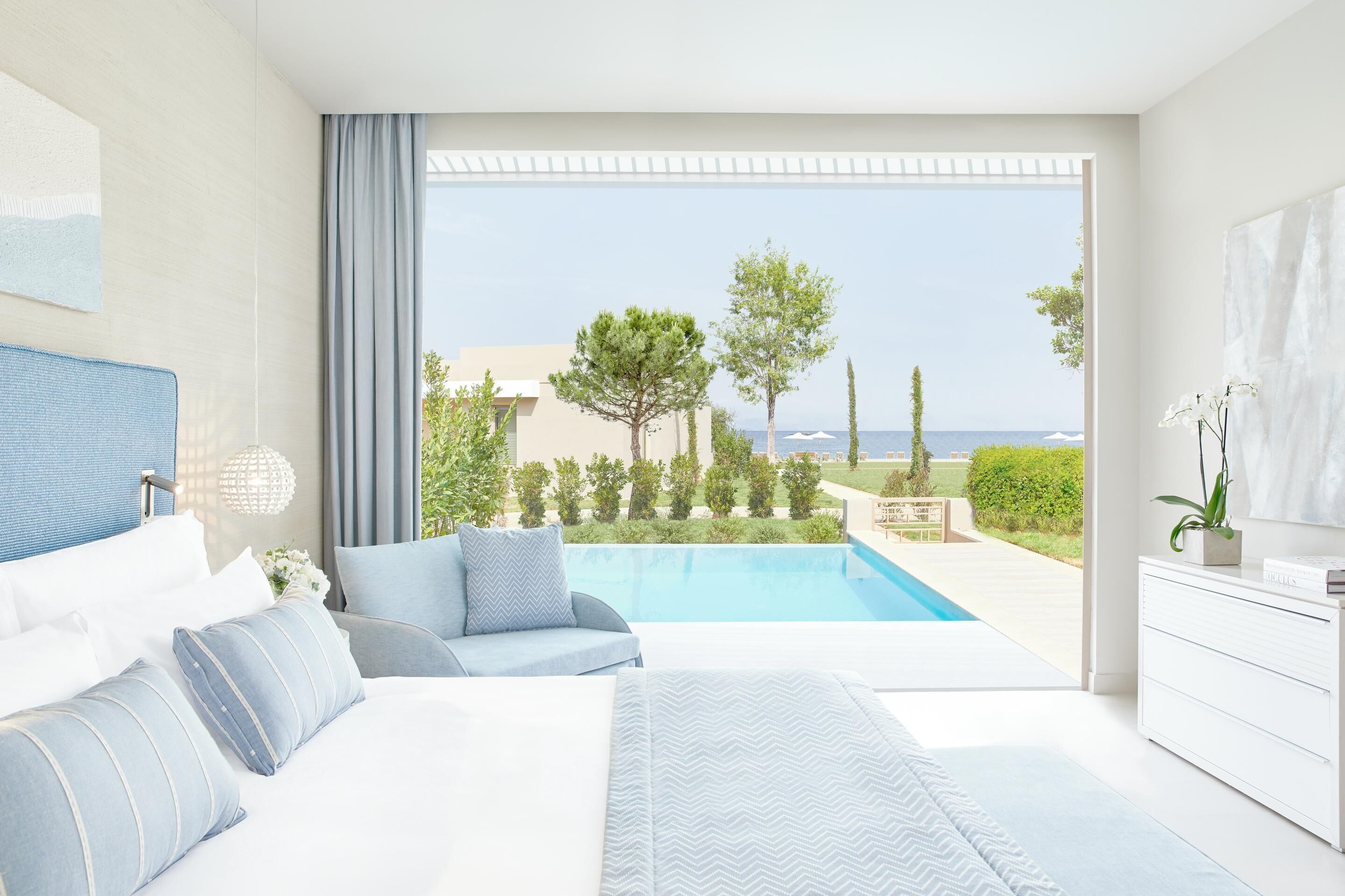 A modern bedroom with a large bed, a seating area, and a view of the sea.