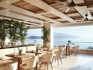 Un restaurante con mesas, sillas, ventanas amplias y plantas en macetas.
